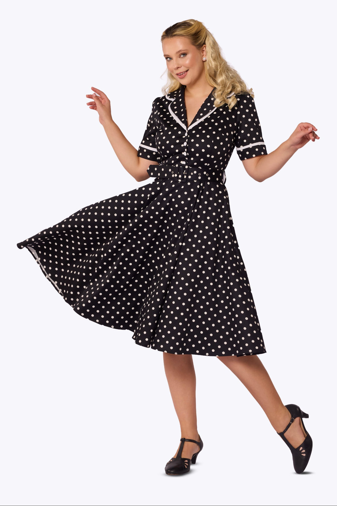 A woman modeling a black and white polka dot midi dress with white trim, posed in a dynamic twirling stance against a plain white studio background.