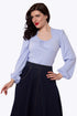 A woman modeling a light blue long-sleeve blouse with a square neckline and gathered front detail, paired with a dark denim skirt, against a plain white studio background.